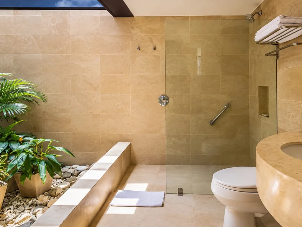 Baño de hotel con ducha de vidrio, lavamanos y acabados en piedra